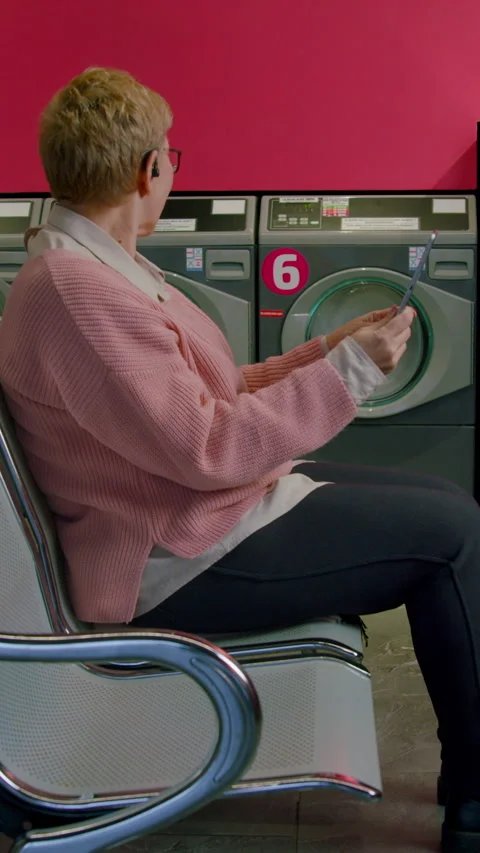 Senior woman multitasking while washing clothes in laundromat, seated on metal Stock Footage 330283176