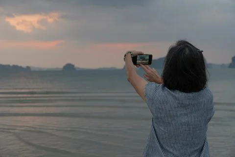 Senior woman using smartphone to take photo of ocean at sunrise during summer Stock Photos