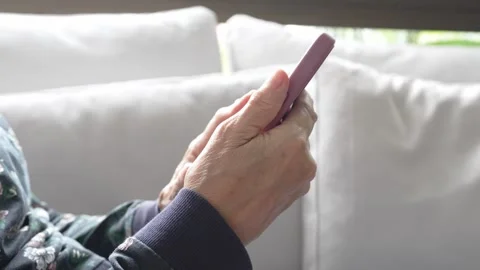 Senior woman using smartphone while relaxing on a gray sofa at home Видео 336337779