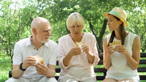Seniors with teenage daughter sitting on bench and eating grilled sandwich with Stock Footage