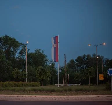 Serbian flag hanging vertically from pole at dusk, illuminated by streetlights Stock Photos