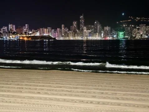 Serene beach perspective looking towards city skyline at night with sand an.. Stock Photos