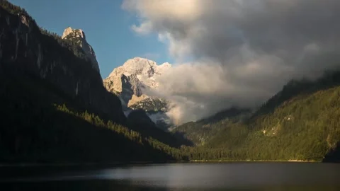 Serene Mountain Lake with Cloud-Covered Peaks and Forested Slopes Reflecting Stock Footage 314788313