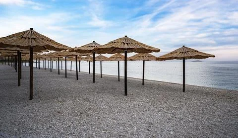 A serene pebble beach lined with rustic straw parasols under a blue sky Stock Photos