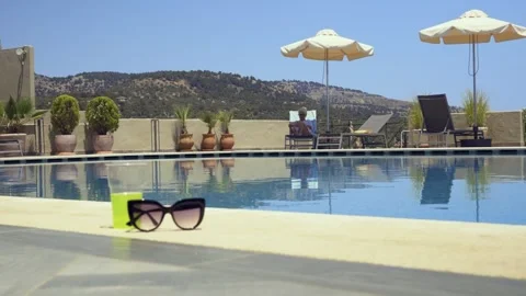A serene poolside setting with sunglasses on the edge, umbrellas and chairs Stock Footage 328658680