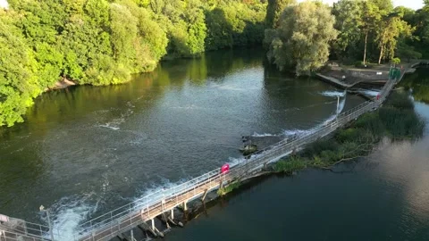 A serene river flows past lush green trees. A walkway spans across the wate.. Stock Footage 340504737