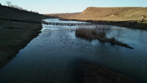 Serene river flows through a grassy landscape with a wooden bridge crossing the Stock Footage 332723625