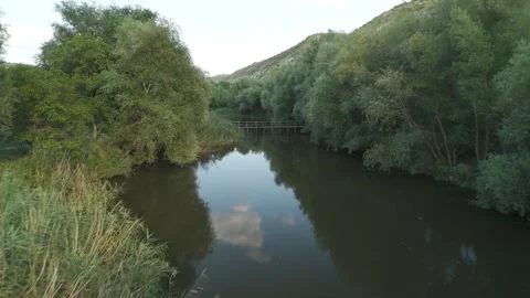 Serene river flows through lush greenery, reflecting trees and clouds, with a Stock Footage 332725676