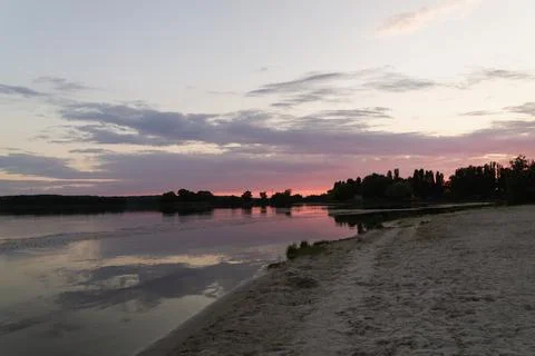 Serene sunset over calm river waters with sandy shore and distant treeline Фото