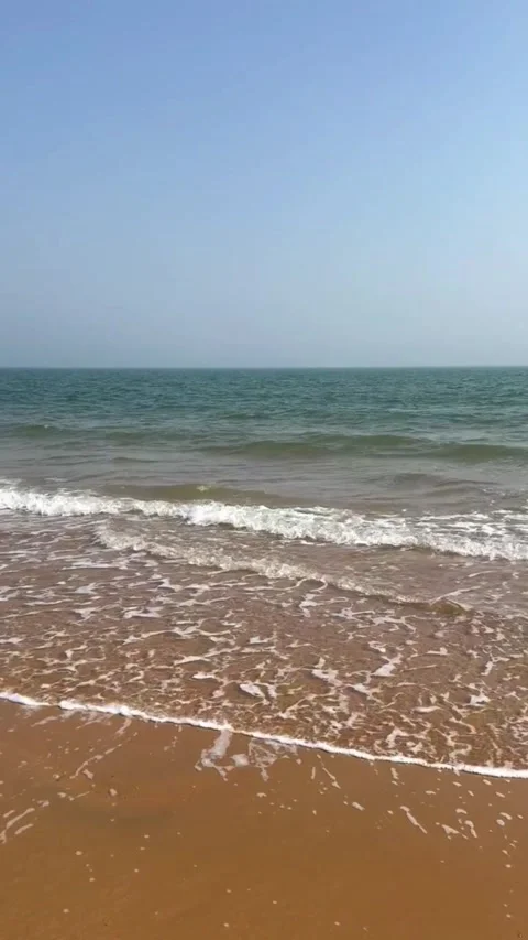 A serene view of gentle ocean waves rolling onto a sandy beach under a clear Vidéo 334163606