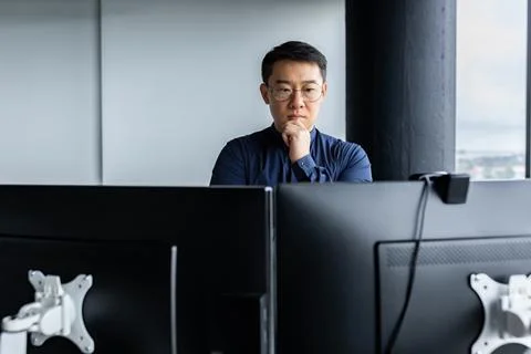 Serious and thinking asian engineer looking at two monitors, close-up photo of Foto stock