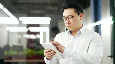 Serious asian businessman is using digital tablet while standing in business off Stock Footage 269387020