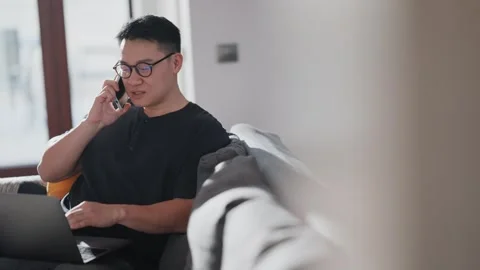 Serious asian man talking on cell phone and working on laptop at home Stock Footage 242007741