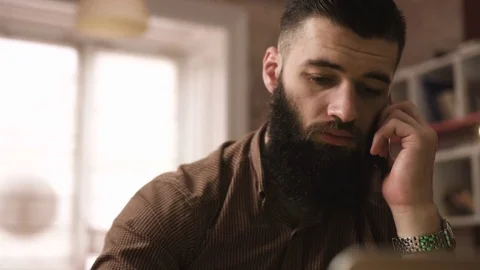 Serious attractive young bearded man sitting at the table and speak the phone in Stock Footage