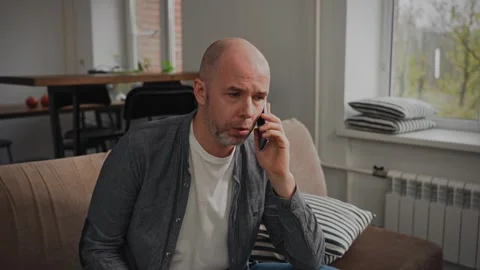 A serious bald middle-aged man sits at home on the sofa, talking to someone on Stock Footage 297050696