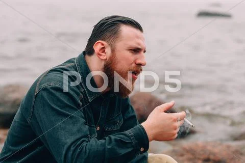 Serious bearded man smoking a cigarette on the beach Stock Photos
