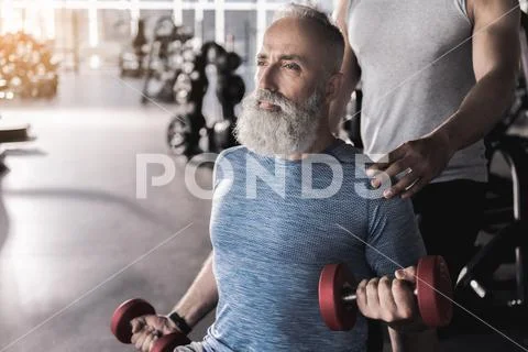 Serious bearded pensioner is doing exercises in athletic center Stock Photos