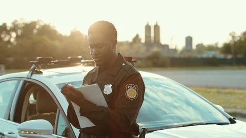Serious black policeman writing notes working outdoors leaning on the police car Video stock 112988190