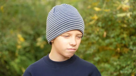 Serious boy in striped hat standing in park looks at camera and nods his head in Stock Footage 169447808