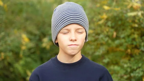 Serious boy in striped hat standing in park looks at camera and nods his head in Stock Footage 169932183