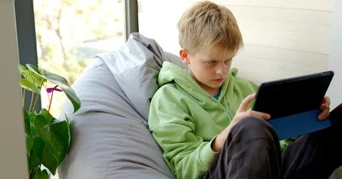 Serious boy using tablet sitting on soft chair, playing online game on tablet 库存照片