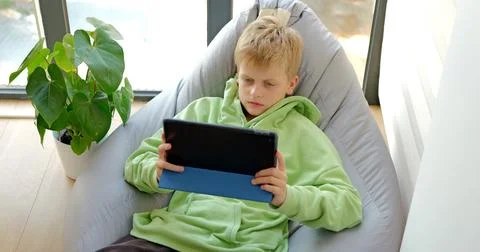 Serious boy using tablet sitting on soft chair, playing online game on tablet 库存照片
