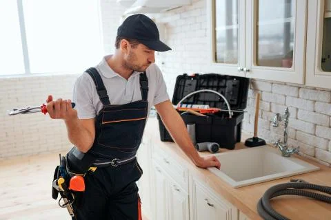 Serious busy young worker lean to sink in kitchen and look at it. He hold wrench Stockfoto's