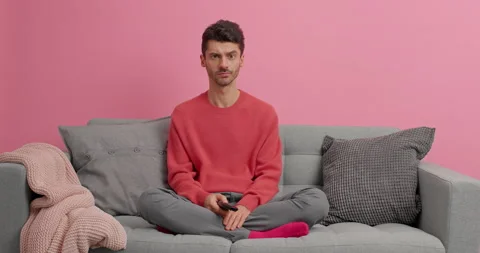 Serious Caucasian man sits on sofa crossed legs switches channels with remote Stock Footage 143634706