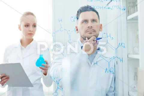 Serious chemist looking at chemical formula on transparent board with his assist Stock Photos