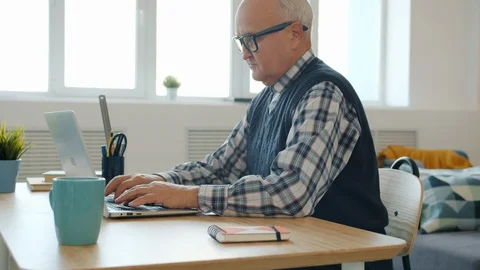 Serious elderly man using computer at home typing sitting at desk indoors Stock Footage 128443635