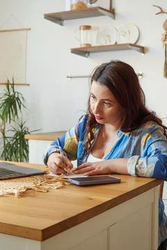 Serious female freelancer making notes on paper while looking at laptop. Remo Stock Photos