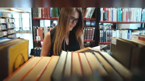 Serious girl student reading a book in the library between the shelves Stock Footage 68141453