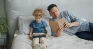 Serious Guy Reading Book In Bed While Small Child Watching Cartoons On Tablet Stock Footage