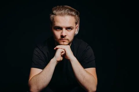 A serious guy sits on black background and leaned on hands, clenched into fist Stock Photos