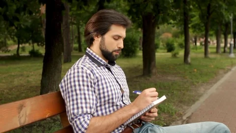 Serious guy is sitting on bench in park and writing notes in notebook. He looks Stock Footage 99739936