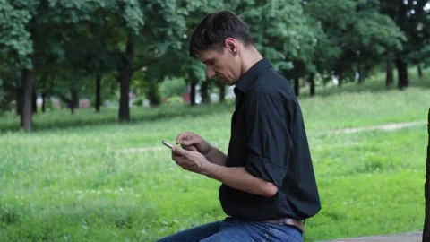 Serious guy sitting in the park chatting via smartphone, blurred background Stock Footage 135238166