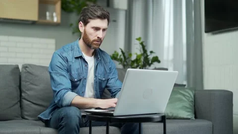 Serious handsome man works remotely at home with laptop while sitting on sofa.  Stock Footage 231233925