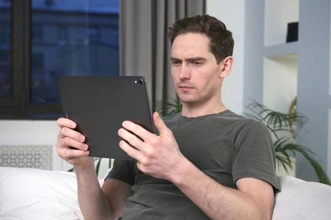 Serious handsome young man is using tablet computer gadget at home, reading or Stock Photos