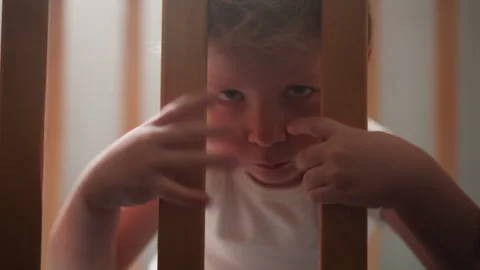 Serious little boy eats snack grabbing wooden bars of crib Stock Footage 243311756