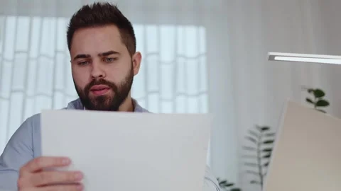 Serious male analysing document hard copy while typing at laptop indoors Stock Footage 262461359