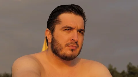 Serious man with bare torso sits looking ahead at sunset Stock Footage 124023989