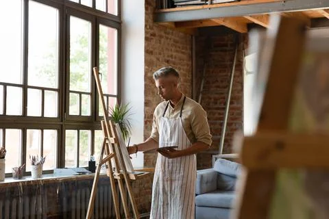 Serious man painter holds palette and paintbrush drawing on canvas Stock Photos