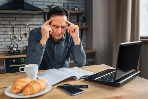 Serious man sit at table in kitchen and reading. He look down at journal and Stock Photos