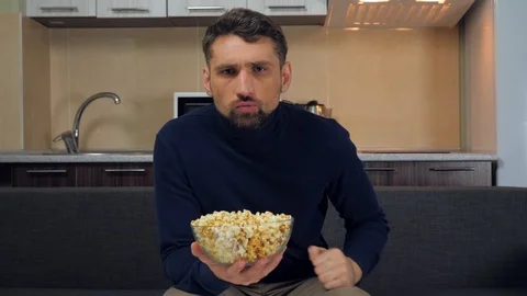 Serious man sitting on the couch, eats popcorn and gets angry, swears on a TV Stock Footage 121840289