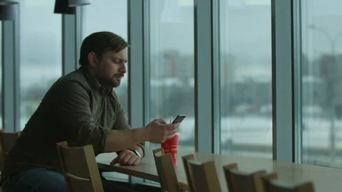 Serious man writes a message and drinks, a large window. Video stock 73340078