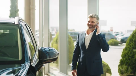 Serious middle age customer standing near new car in auto showroom. Making his Stock Footage 240193243