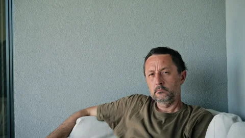 Serious middle-aged man sits on a white sofa against a neutral gray wall, a.. Stock Footage 335825593