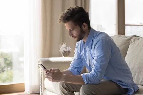 Serious millennial man using cellphone seated on sofa at home 写真素材