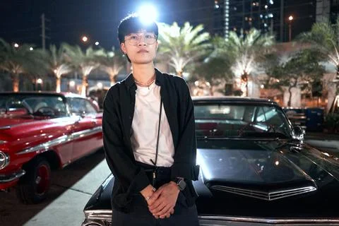 Serious non-binary person with glasses leaning on a car in an urban night fair Stock Photos