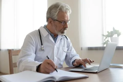 Serious senior physician using computer app making notes in journal Stock Photos